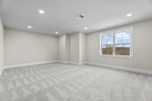 Empty room with beige walls, light gray carpet, a rectangular window showing a blue sky, and recessed ceiling lights.