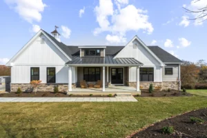 A modern white house with a gabled roof and stone accents, featuring a covered porch with wooden chairs, surrounded by a landscaped yard.