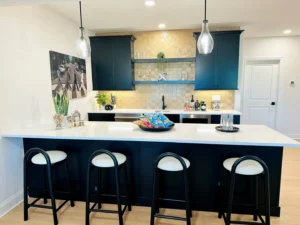 Modern kitchen with black cabinets, pendant lights, and a white island with four black stools. Yellow tile backsplash, plants, and decorative items add accents.