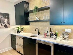 Modern black kitchen with a white countertop, bar essentials, glassware, plant, and wall art. Upper cabinets and open shelves are mounted on a geometric tiled backsplash.