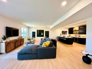 Spacious open-concept living area with a sectional sofa, wall-mounted TV, guitars on display, and a kitchen with a bar in the background. Light wood flooring and modern decor elements.