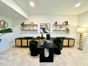 Modern home office with a black desk, two green chairs, and shelves with decor. Neutral tones and abstract art enhance the space. Floor lamp with stacked design on the right.