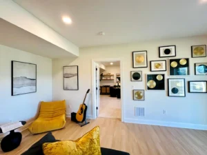 Room with yellow seating, a guitar, and wall art featuring multiple framed records. Doorway leads to another room with shelving. Light wooden floor and neutral walls.