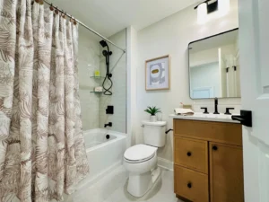 A bathroom with a patterned shower curtain, a bathtub, a toilet, a wooden vanity with a sink, and a mirror above. A framed picture and a small plant are on the wall.