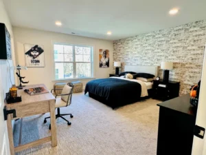 Bedroom with a large bed, black and white bedding, desk with a chair, patterned accent wall, two photos, window with tree view, and neutral carpeting.