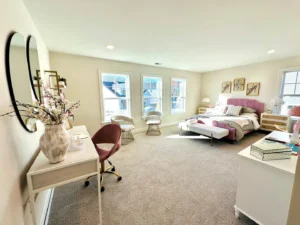 Bright bedroom with a neatly made bed, two chairs by large windows, a desk with a vase and mirror, and neutral decor.