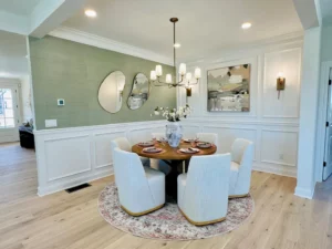 Dining room with a round table, white upholstered chairs, chandelier, and decorative wall art. Green accent wall and light wood flooring create a modern, elegant atmosphere.