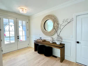 Bright entryway with a wooden table, round mirror, and decorative vases. Double glass doors are on the left, and a closed door is on the right. Light wood flooring and white walls.