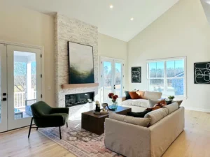 Modern living room with high ceilings, a stone fireplace, two couches, a green chair, and large windows. Neutral colors with abstract artwork and a patterned rug.