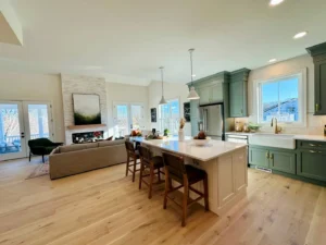 Open-concept kitchen and living area with hardwood floors, large island, modern green cabinets, pendant lights, and cozy seating by a fireplace. Sunlight streams in through large windows.