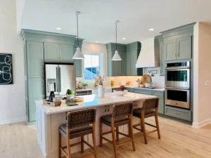 Modern kitchen with light green cabinets, stainless steel appliances, and a large island with three woven chairs. White countertops and pendant lights complete the bright, airy space.