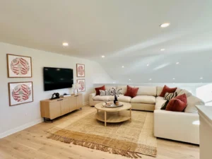 A modern living room with a white sectional sofa, red and beige pillows, a round wooden coffee table, a wall-mounted TV, and framed art on white walls. Natural light illuminates the space.