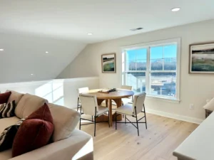 Bright room with round wooden table and chess set by a window. Beige couch with red cushions on the left, and framed landscape paintings on the walls. Light wood floor.