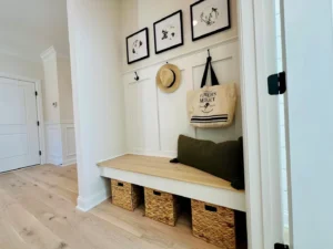 Mudroom with a wooden bench, three woven baskets underneath, and hooks holding a tan hat and tote bag. Above are three framed nature illustrations. Light wood floor and white walls.