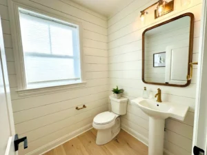 A bright bathroom with a window, toilet, and pedestal sink. A rectangular mirror with lights above it and a potted plant are on the wall. The room has shiplap walls and light wood flooring.