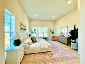 Spacious bedroom with a large bed, white furniture, pink armchair, dresser, green plants, and artwork. Natural light from big windows illuminates the room.