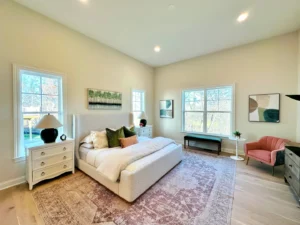 A spacious bedroom with a large bed, white walls, colorful artwork, a beige rug, and large windows. There's a pink chair, a black bench, and two nightstands with lamps.