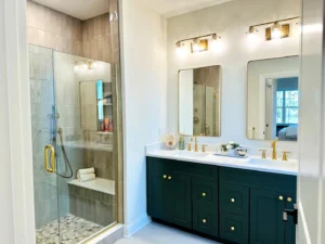 Modern bathroom with dual sinks, gold fixtures, and a dark green vanity. A glass-enclosed shower is on the left. Neutral tiles and soft lighting provide a clean, contemporary look.